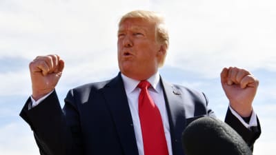 Donald Trump reagerar genom att knyta bägge händer. Han är fotad utomhus, iklädd mörkblå kostym, vit skjorta och röd slips. 