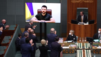 Volodymyr Zelenskyj på en tv-skärm framför ett rum med parlamentsledamöter i Australien. De klappar i händerna.