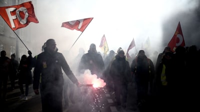 Demonstranter med röda flaggor med bokstäverna FO i vit text. Det är flera människor på rad och bilden är mörk och det är rökigt.