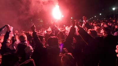 Hela bilden fylls av fans, vissa har händerna upp i luften och rök gör att bilden färgas röd och svart.