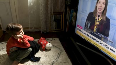 Litet barn sitter på golvet i hemmiljö, tittar på Li Andersson i regeringens coronainfo på tv.