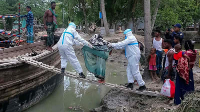Två hjälparbetare iklädda skyddsutrustning mot coronaviruset hälper en person att ta om bord sig på en båt. De har en spång från flodbanken till båten.