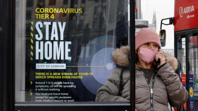 En kvinna med munskydd på sig i London. Hon står bredvid en skylt som uppmanar britterna att stanna hemma och följa de rådande coronarestriktionerna.