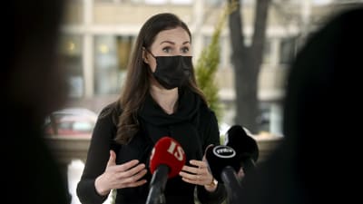 Svartklädd kvinna i svart munskydd talar utomhus med många mikrofoner framför sig. Statsminister Sanna Marin (SDP) kommenterar träffen med riksdagsgrupperna den 25 februari 2021.