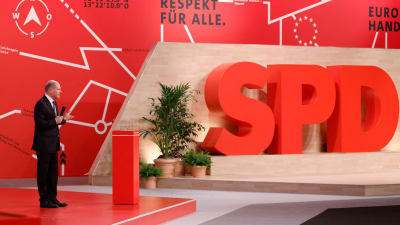 Olaf scholz står på ett podium - i bakgrunden syns stora bokstäver i rött som bokstaverar SPD