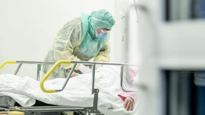 Person iklädd skyddmundering tar blodprov av person som ligger på en sjukhussäng.