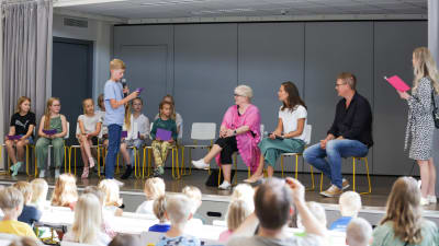 Riksdagsledamot Tuula Väätäinen, statsminister Sanna Marin joch SDP:s riksdagsgrupps ordförande Antti Lindtman sitter på en scen med skolbarn. ett barn har stigit upp och ställer en fråga.