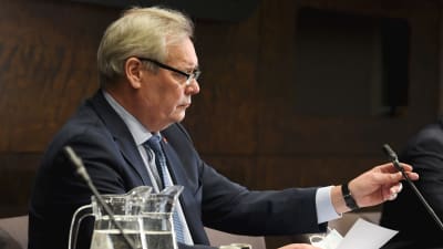 En glasögonprydd och kostymklädd Antti Rinne sitter i profil framför en mikrofon.