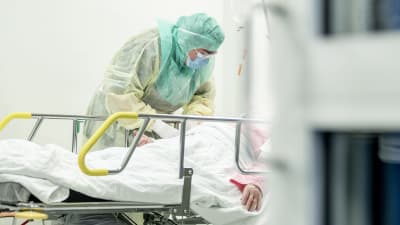 Vårdare i skyddsutrustning tar hand om en patient som ligger i en sjukhussäng.