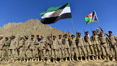 Ett tjugotal krigare i khakiuniform står uppstållda på rad och viftar med motståndsrörelsens grön-vit-svarta och Afghanistans nationella grön-röd-svarta flagga.