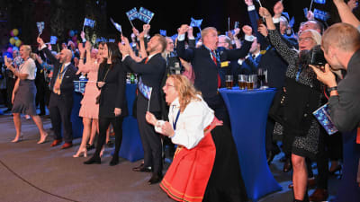 Anhängare av Sverigedemokraterna jublar på valvaka, många håller i blåa flaggor och i förgrunden en kvinna klädd i folkdräkt.