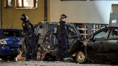 Två män i polisuniformer står på en gata eller en gård framför bilar skadade av en explosion.  Kriminaltekniker arbetar på platsen efter den kraftig explosionen som inträffade i under natten mot måndagen på Östermalm i Stockholm den 13 januari 2020..
