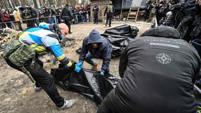 Poliisit ja kaupungin työntekijät kantavat kuusi osittain palanutta ruumista ruumispusseihin, toimittajat kuvaavat taustalla. 