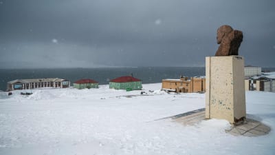En byst på en piedestal föreställer en man som tittar ned på en samling hus vid vattnet. Snö ligger på marken.