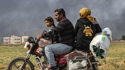 En syrisk familj flydde från staden Ras al-Ain på en motorcykel på söndagen. Röken bakom dem kommer från brinnande bildäck. Man har satt eld på dem för att skymma sikten för turkiskt krigsflyg. 