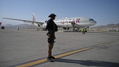 En soldat står på ett flygfält med ett passagerarplan bakom sig. På planet står med stora bokstäver Qatar.