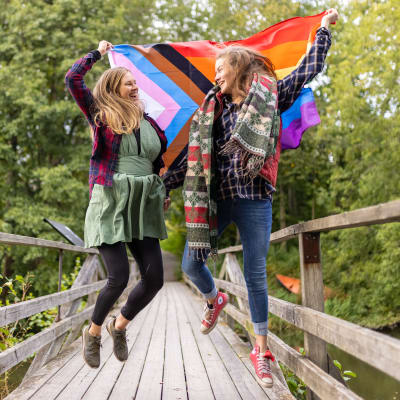 Två kvinnor håller i en flagga i många färger och hoppar upp i luften på en mindre gångbro.