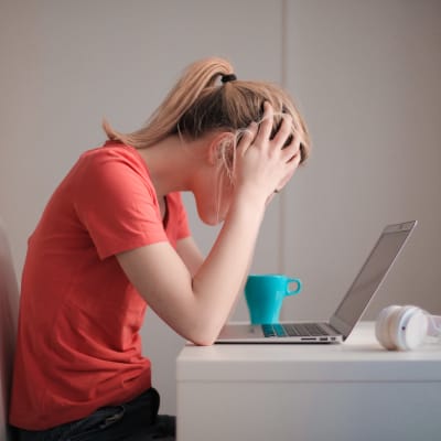 Kvinna i röd t-shirt sitter vid ett bord med uppfälld laptop och lutar huvudet i händerna 