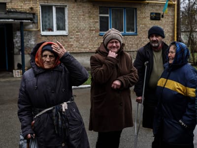 Tre älde kvinnor och en äldre man på kryckor står och gråter framför en byggnad. I bakgrunden skymtar en ukrainsk flagga.