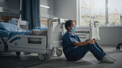 En sjukskötare i munskydd sitter på golvet vid en sjukhussäng.