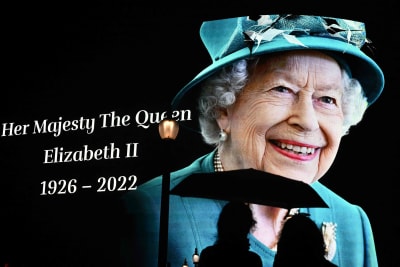 En bild på drottning Elizabeth den andra på en stor skärm i London. I förgrunden syns två personer som står och ser på bilden.