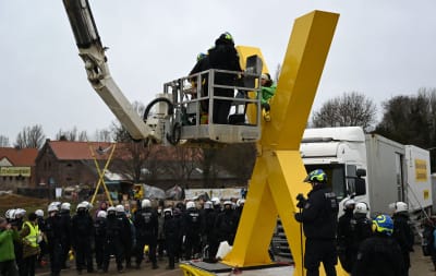 Polisen står i en lyftkranskorg och förbereder sig på att lyfta iväg en demonstrant som sitter på en stor x-formad skulptur.