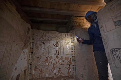 En man tittar in i en gravkammare med egyptiska bilder på väggarna. I sin hand håller mannen en mobiltelefon och lyser upp bilderna på en vägg.