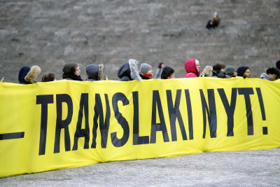 Demonstranter som bär upp en stor gul flagga med texten Translagen nu! 