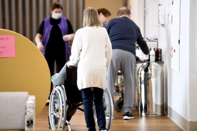 Vy från ett äldreboende, med en person som rullar fram en annan i rullstol.