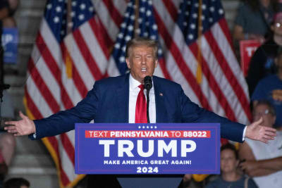 Donald Trump står vid ett talarpodium med armarna utsträckta och talar i en mikrofon. Han är klädd i blå kavaj och röd slips. Bakom honom hänger amerikanska flaggor och på podiet står det Trump och Make America Great Again! 2024