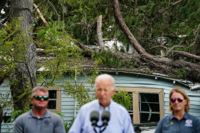 En suddig Joe Biden i förgrunden, i bakgrunden träd som fallit över ett hus.