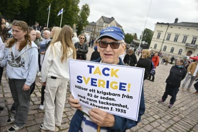 En äldre man i blå keps står på torget i Lovisa och håller i en skylt med texten "Tack Sverige! Hälsar ett krigsbarn från 82 år sedan. Med varm tackamhet, Pauli Ahvonen f. 1935".