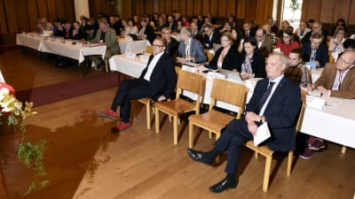 SDP:s Antti Rinne sitter på en stol och lyssnar med många andra människor.