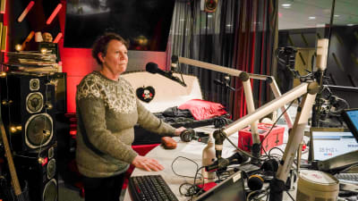 En kvinna i stickad tröja står i en radiostudio. Hon lutar sig mot ett bord och har en mikrofon framför sig.