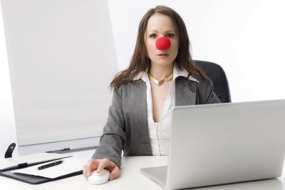 Nainen istuu tietokoneen ääressä punainen leikkinenä päässään ja kameraan katsoen.