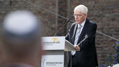 En man i kostym står vid ett podium. Han har vit skjorta och randig slips. Han ser allvarlig ut och han vitt hår och glasögon. I förgrunden till vänster ett huvud som är iklätt en kippa.