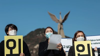 Tre kvinnor med munskydd håller upp plakat med koranska tecken. I bakgrunden en staty av Fågel Fenix.