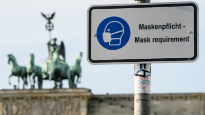 På en vit skylt står det att munskydd krävs - en bild av en person klädd i munskydd finns på skylten i blått. I bakgrunden syns ett monument där fyra hästar drar en vagn med en person i.
