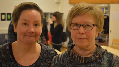 Två damer med mörkblommiga skjortor, den till vänster har mörkt hår, den till höger ljust hår och glasögon. De står i en sal med bord och mötespapper.