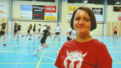 En kvinna i röd t-skjorta i förgrunden. I bakgrunden handbollsspelare mitt i en match.