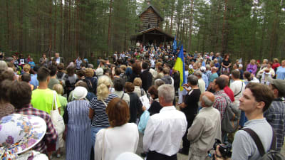 Hundratals människor står samlade i skogen framför ett stockhus på en kulle.