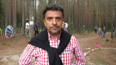 Aleksandr Arkangelskij, en mrkårig medelålders man med skägg och röd-vit-rutig skjorta stirrar stint in i kameran med skog och människor i bakgrunden.