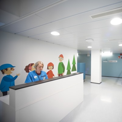 En korridor med en disk. Bakom disken sitter en kvinna med hälsocentralskläder.
