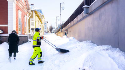 En man i neonfärgad arbetsdräkt skyfflar snö vid kanten av den slätplogade trottoaren, en person med ryggen åt kameran går längs trottoaren, vädret är soligt.