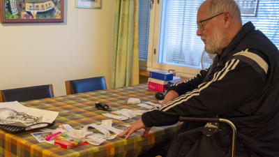 En äldre man sitter på sin rullator vid ett bord, där det finns en mängd kvitton.