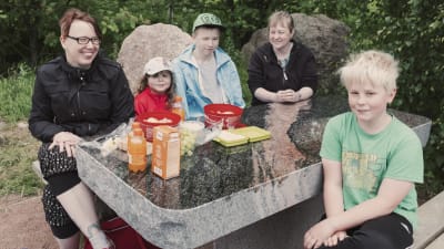 Två vuxna och tre barn har picknick kring ett bord gjort av polerad granit.