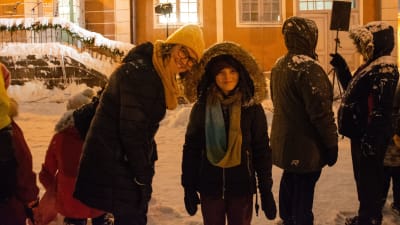 En kvinna lutar sig närmare ett barn. Båda ler och tittar in i kameran. De står i snöyran framför Gamla rådhuset.