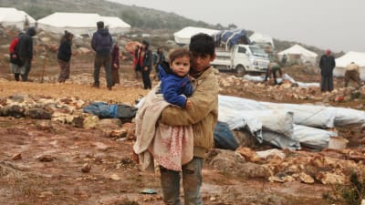 Bild på äldre barn som håller yngre barn i famnen. I bakgrunden syns flyktingläger under konstruktion.