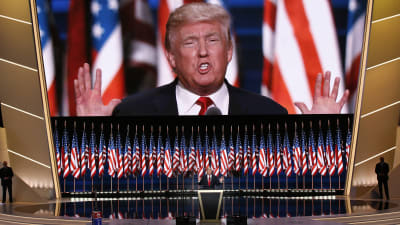 Donald Trump står bakom ett guld- och svartfärgat podium med flera amerikasnka flaggor och en enorm screen bakom honom som streamar talet den 21 juli 2016.
