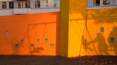 En orange husvägg med skuggorna från barn som gungar och vuxna som ger fart i en lekpark.